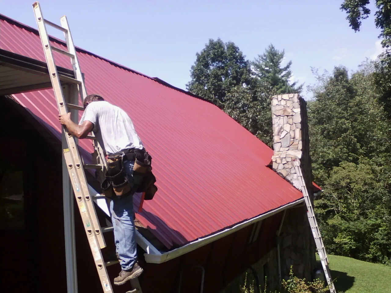 Roofer on a ladder beside a red metal roof and stone chimney — Soffit & Fascia Repair by Roofing Brothers in Jersey
